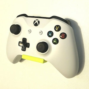 May include: A white Xbox controller with black buttons and a yellow accent. The controller is mounted on a wall.