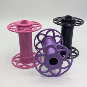 May include: Three cylindrical spools for thread or yarn, each featuring a central column and circular end caps with a decorative cutout pattern. One spool is light pink, one is purple, and one is black, all standing upright on a flat surface.