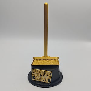May include: A gold-colored trophy shaped like a broom. The broom is mounted on a black base with the text "NUMBER ONE SODOR STATION SWEEPER" in gold lettering. The trophy is set against a white background.