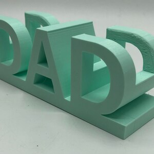 May include: A three-dimensional, light mint green plastic sign featuring the word DAD in bold, block capital letters. The object is constructed with a flat base and a layered, textured surface finish consistent with a 3D printed manufacturing process.