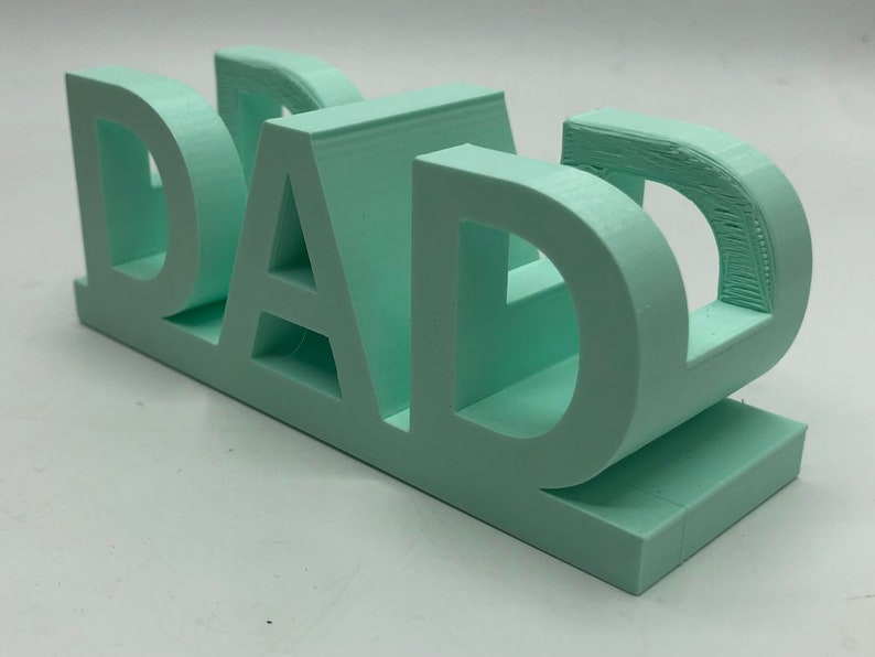 May include: A three-dimensional, light mint green plastic sign featuring the word DAD in bold, block capital letters. The object is constructed with a flat base and a layered, textured surface finish consistent with a 3D printed manufacturing process.
