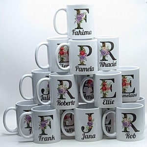 May include: A stack of 12 white ceramic mugs with floral designs and a letter of the alphabet on each mug. The mugs are arranged in a pyramid shape. The names on the mugs are Fahima, Pamela, Rhaciel, Juli, Roberto, Ellie, Frank, Jana, and Rob.