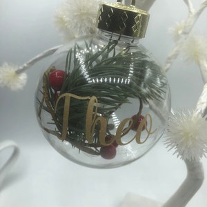 May include: A clear spherical Christmas ornament featuring a gold cap and a gold string. Inside the transparent globe are green pine needles and small red berries. The name Theo is written on the front of the ornament in gold cursive lettering.
