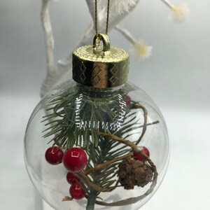 May include: A clear spherical Christmas ornament featuring a gold-colored cap and a gold hanging string. Inside the transparent globe are green pine needles, small red berries, a brown pinecone, and thin brown twigs arranged decoratively.