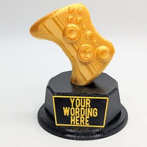May include: A gold-colored video game controller trophy with a black base. The base has a black plaque with the text "YOUR WORDING HERE" in yellow.