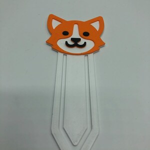 May include: Orange and white bookmark with a cartoon dog face. The bookmark has a white, three-pronged design.