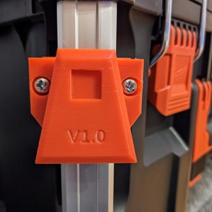 May include: Close-up of an orange plastic latch with the text "V1.0" on it. The latch is attached to a silver metal bar.
