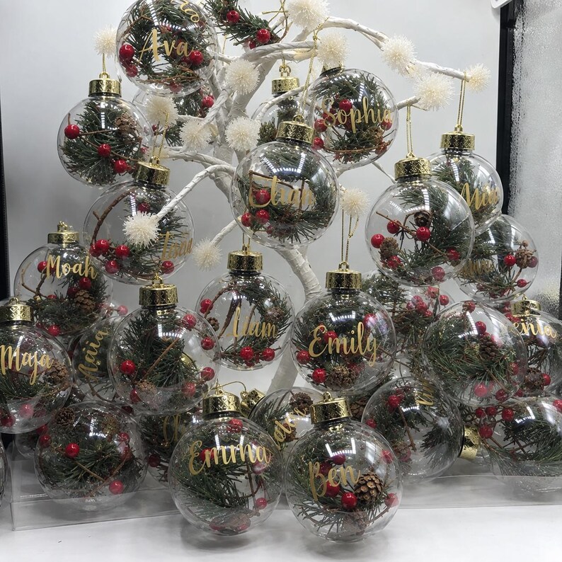 May include: A collection of clear, spherical Christmas ornaments hanging from a white decorative tree. Each ornament contains green pine needles, small brown pinecones, and red berries, with a gold cap and a custom name written in gold script.