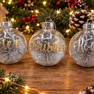 May include: Three clear spherical glass ornaments with silver tinsel filling. Each ornament features a name in cursive lettering: Peter and Christin in gold, and Roosy in silver. Each has a silver metal cap and a hanging loop.