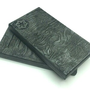May include: Two rectangular dark grey blocks featuring a textured, wavy pattern resembling wood grain or layered metal. The top block displays a circular emblem with a six-spoked star design in the upper left corner, embossed on the surface.