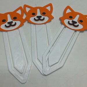 May include: Three white bookmark holders with orange and black cartoon corgi dog designs. The bookmarks are made of a thick, sturdy material and have a slightly textured surface.