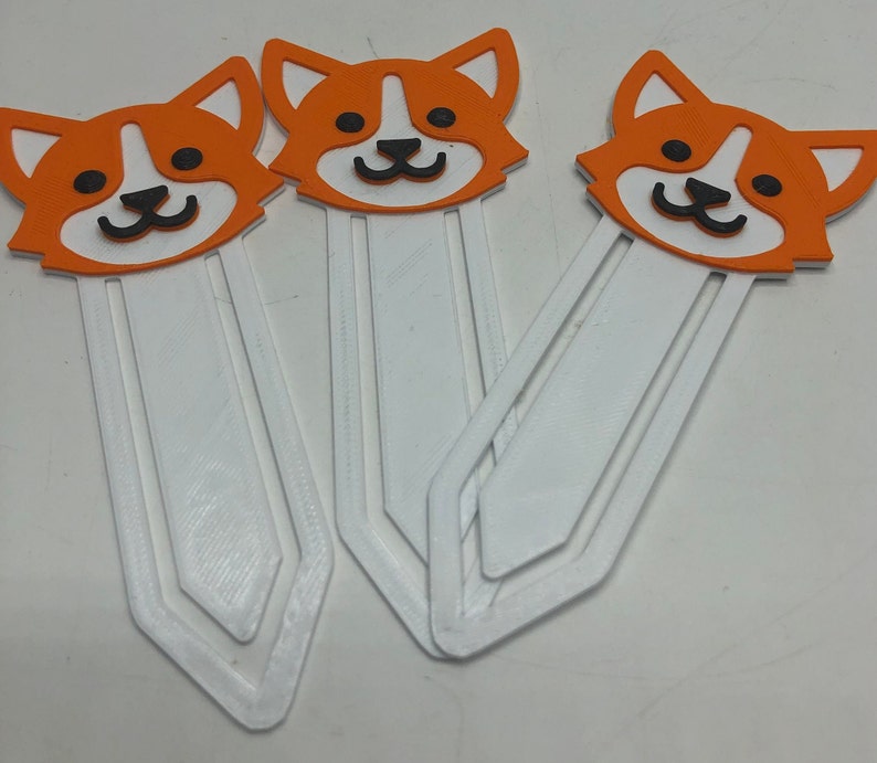 May include: Three white bookmark holders with orange and black cartoon corgi dog designs. The bookmarks are made of a thick, sturdy material and have a slightly textured surface.