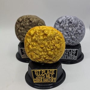 May include: Three cookie-shaped trophies on black hexagonal bases. The front trophy is gold with a plaque reading 1ST PLACE 2025 MLC COOKIE BAKE-OFF. The back left is bronze, and the back right is silver, both with corresponding place plaques.