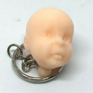 May include: A keychain featuring a small, realistic-looking baby head made of a smooth, light pink material. The head is attached to a silver metal key ring.