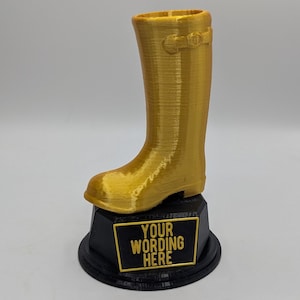 May include: A gold-coloured boot trophy on a black base. The boot features a buckle detail and is mounted on a black base with the text "YOUR WORDING HERE" in gold lettering.