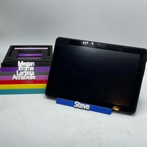 May include: A black tablet computer with a blue stand that says "Steve". The tablet is sitting on top of a stack of colourful, rectangular blocks with the names "Megan", "Emma", "Larissa", and "Annabelle" printed on them.