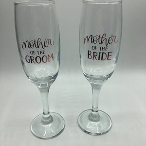 May include: Two clear glass champagne flutes with gold lettering. The left flute says "mother of the groom" and the right flute says "mother of the bride".