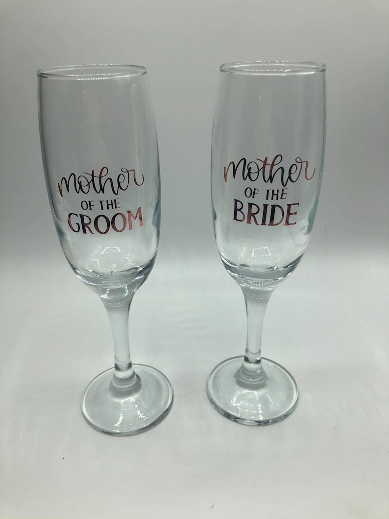 May include: Two clear glass champagne flutes with gold lettering. The left flute says "mother of the groom" and the right flute says "mother of the bride".