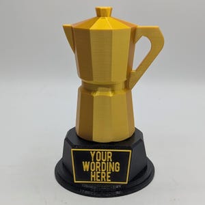 May include: A gold-colored coffee maker trophy with a black base. The base has a rectangular plaque with the words "YOUR WORDING HERE" in gold lettering. The coffee maker is a detailed, geometric design.