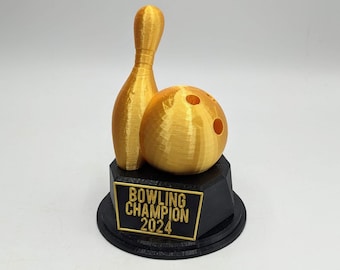 May include: A gold-colored 3D printed bowling pin and bowling ball trophy mounted on a black hexagonal base. The base features a rectangular plaque with the gold-colored text "BOWLING CHAMPION 2024" centered on the front face of the trophy.