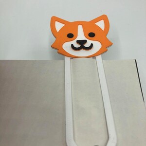 May include: Orange and white cartoon dog bookmark with a white metal clip. The dog has black eyes and a black nose.