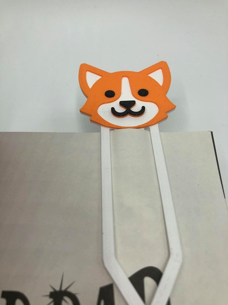May include: Orange and white cartoon dog bookmark with a white metal clip. The dog has black eyes and a black nose.