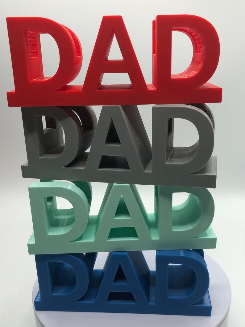 May include: A stack of four three-dimensional block signs, each featuring the word DAD in capital letters. The top sign is red, the second is gray, the third is light green, and the bottom sign is blue. Each sign has a flat base and top.