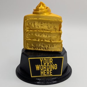 May include: A gold-colored cake trophy with a black base. The base has a black plaque with the text "YOUR WORDING HERE".