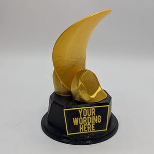 May include: A gold-colored trophy with a curved, wing-like element and a rounded shape, set on a black base. The base features a rectangular plaque with the text "YOUR WORDING HERE" in gold.