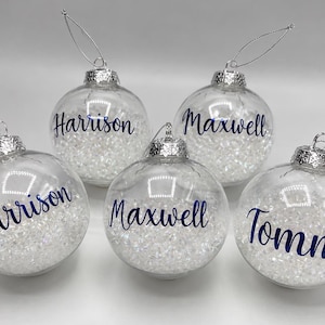 May include: Five clear, spherical glass ornaments filled with small, white, crystalline beads. Each ornament features a silver cap with a hanging string and is personalized with dark blue script text reading Harrison, Maxwell, or Tommy.