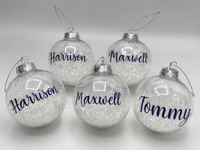 May include: Five clear, spherical glass ornaments filled with small, white, crystalline beads. Each ornament features a silver cap with a hanging string and is personalized with dark blue script text reading Harrison, Maxwell, or Tommy.