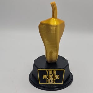 May include: A gold-colored trophy in the shape of a chili pepper, mounted on a black base. The base has a rectangular plaque with the words "YOUR WORDING HERE" in gold lettering.