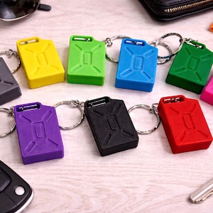 May include: A collection of colorful, miniature jerry can keychains in various colors including gray, yellow, green, blue, green, pink, purple, black, and red. Each keychain has a silver keyring attached. A car key and sunglasses are also visible.