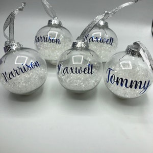 May include: Four clear glass baubles with silver ribbon and white glitter inside. The baubles are personalised with the names "Harrison", "Maxwell", and "Tommy".
