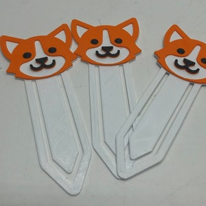 May include: Three orange and white 3D printed bookmark designs featuring a cartoon dog with black eyes and a black nose. The bookmarks are white with a white tab at the top.