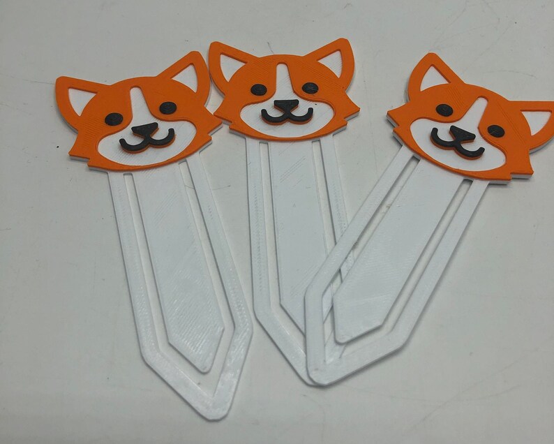 May include: Three orange and white 3D printed bookmark designs featuring a cartoon dog with black eyes and a black nose. The bookmarks are white with a white tab at the top.