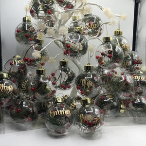 May include: A collection of clear spherical glass ornaments hanging from a white decorative tree. Each ornament contains green pine needles, small brown pinecones, and red berries, with gold cursive names printed on the exterior surface.