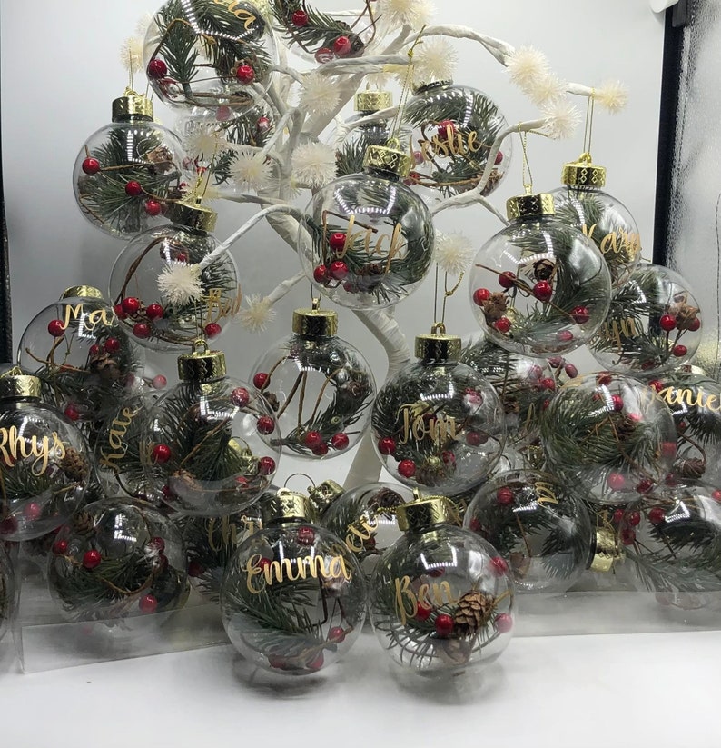 May include: A collection of clear spherical glass ornaments hanging from a white decorative tree. Each ornament contains green pine needles, small brown pinecones, and red berries, with gold cursive names printed on the exterior surface.