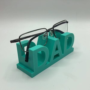 May include: A teal-colored 3D printed glasses stand shaped like the word DAD, featuring a pair of dark-rimmed eyeglasses with the text PURE TITANIUM 53 17-140 CE printed on the inner temple, resting securely on the top of the letters.