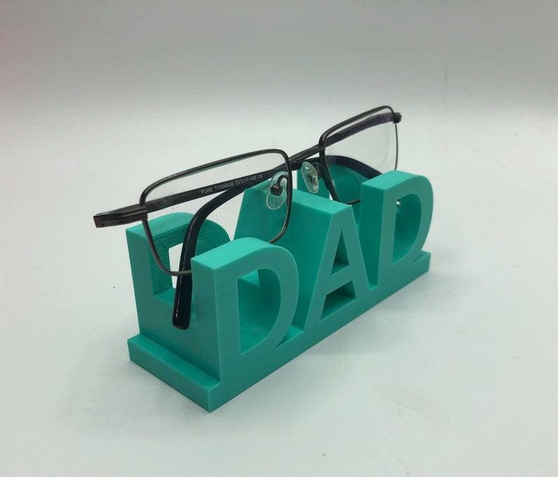May include: A teal-colored 3D printed glasses stand shaped like the word DAD, featuring a pair of dark-rimmed eyeglasses with the text PURE TITANIUM 53 17-140 CE printed on the inner temple, resting securely on the top of the letters.
