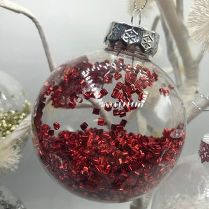 May include: A clear spherical glass ornament filled with small, metallic red rectangular confetti pieces. The ornament features a silver-colored metal cap with a snowflake pattern and a thin silver hanging string attached to the top.
