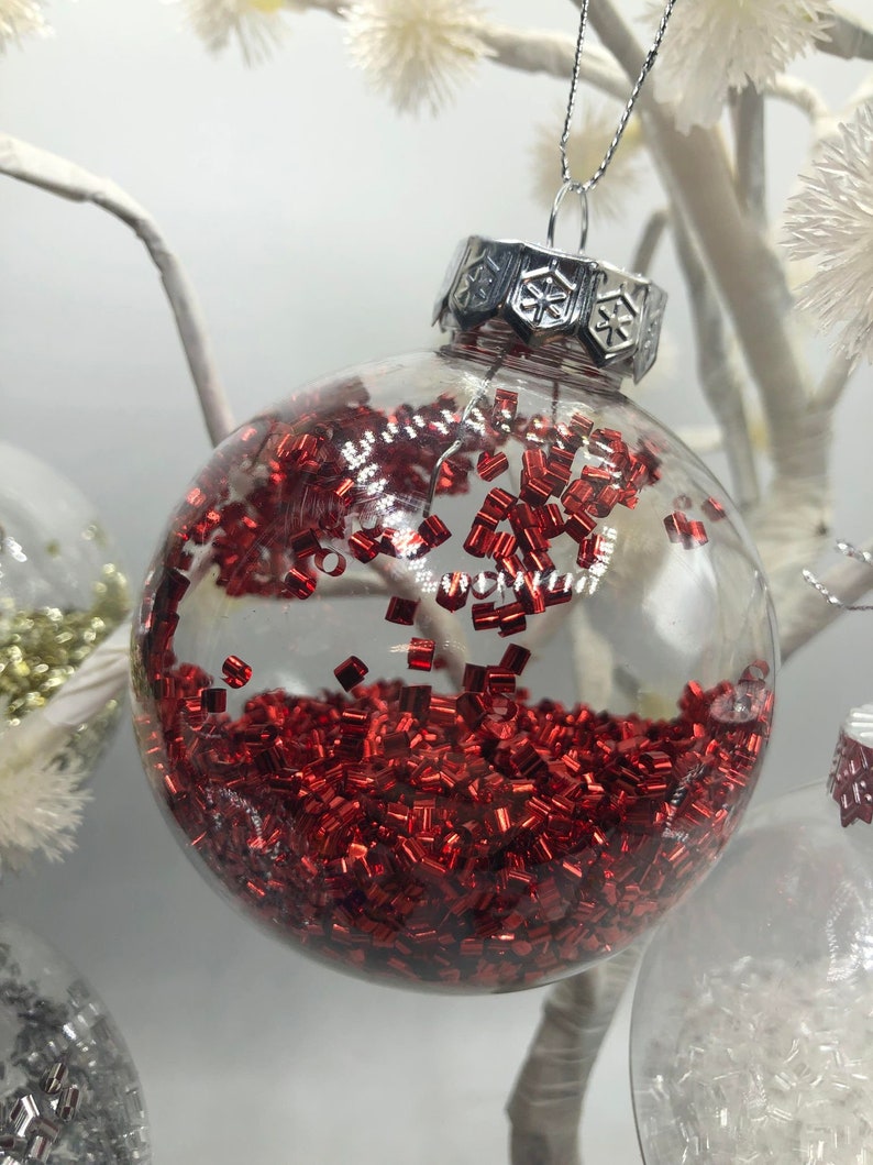 May include: A clear spherical glass ornament filled with small, metallic red rectangular confetti pieces. The ornament features a silver-colored metal cap with a snowflake pattern and a thin silver hanging string attached to the top.