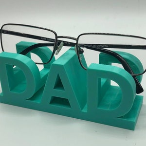 May include: A teal-colored 3D printed glasses stand shaped like the word DAD, featuring a block-letter design. A pair of black-framed eyeglasses with the text PURE TITANIUM 53-18-140 CE and GUNMETAL printed on the inner temple rests on the stand.