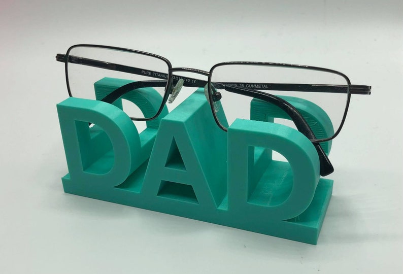 May include: A teal-colored 3D printed glasses stand shaped like the word DAD, featuring a block-letter design. A pair of black-framed eyeglasses with the text PURE TITANIUM 53-18-140 CE and GUNMETAL printed on the inner temple rests on the stand.
