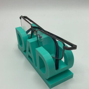 May include: A teal-colored 3D printed eyeglass stand shaped like the word DAD, featuring a flat base and upright letters that hold a pair of dark gunmetal-colored rectangular eyeglasses with clear lenses resting across the top of the letters.