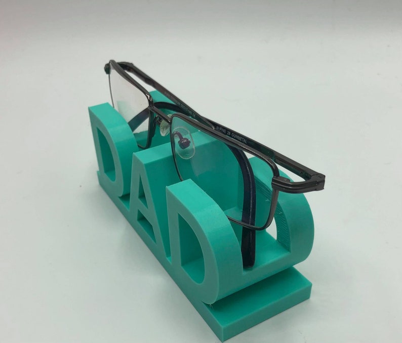 May include: A teal-colored 3D printed eyeglass stand shaped like the word DAD, featuring a flat base and upright letters that hold a pair of dark gunmetal-colored rectangular eyeglasses with clear lenses resting across the top of the letters.