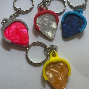 May include: Four guitar pick keychains with different colored plastic frames and a variety of patterned guitar picks. The picks are white, pink, blue, and gold.