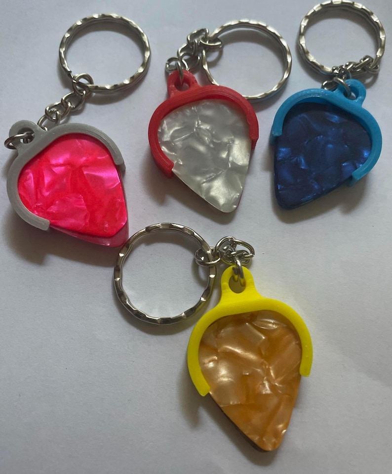 May include: Four guitar pick keychains with different colored plastic frames and a variety of patterned guitar picks. The picks are white, pink, blue, and gold.