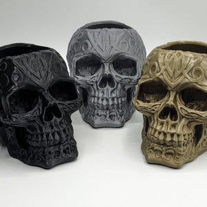 May include: Three decorative skull planters in black, grey, and brown. Each skull has intricate carvings and a detailed design.
