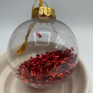 May include: A clear, spherical glass ornament filled with small, metallic red cylindrical beads. The ornament features a gold-colored metal cap with a geometric star pattern and a yellow ribbon loop for hanging.